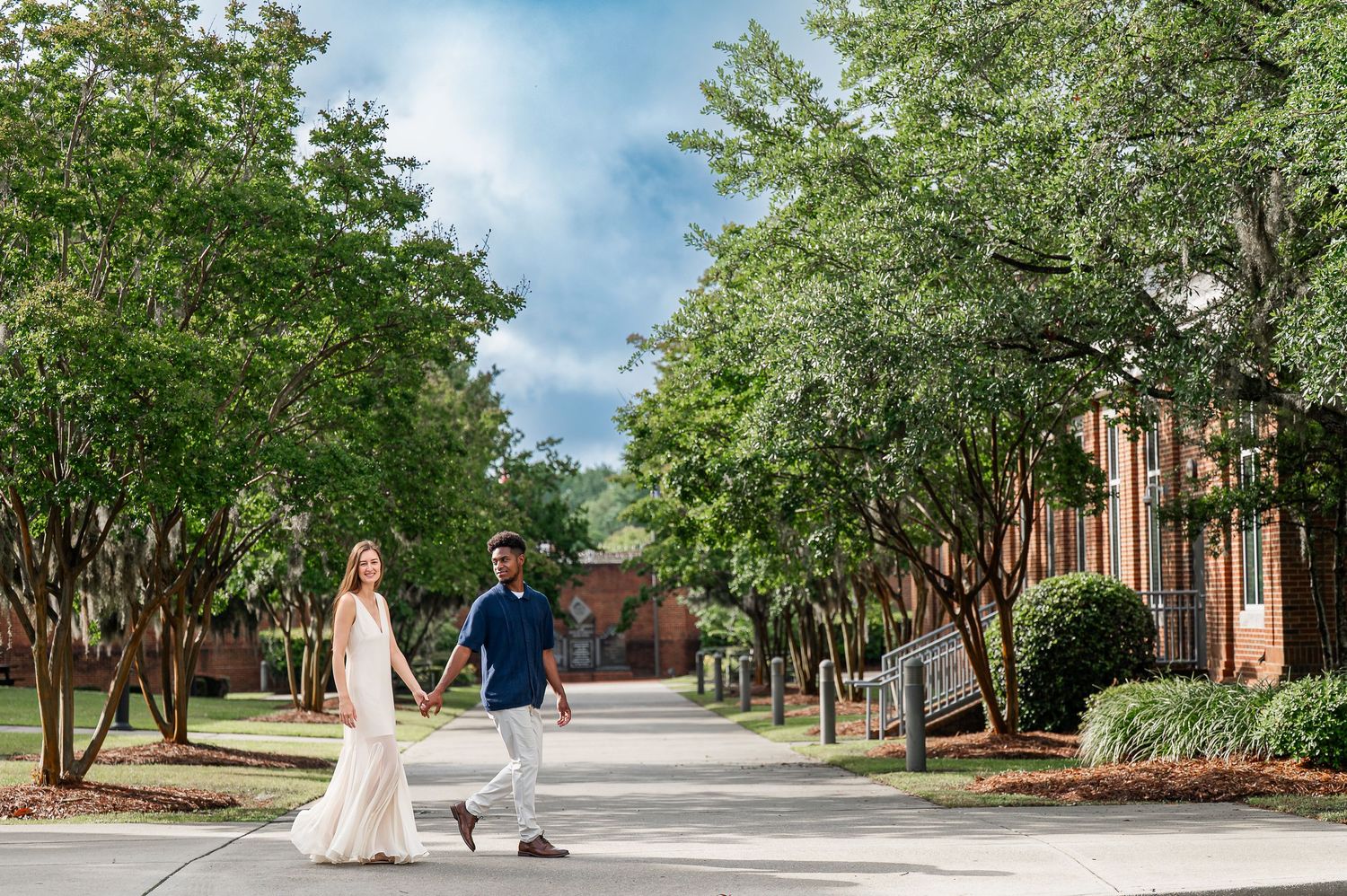 Wide landscape view of a couple walking along a tree-lined path near brick buildings.