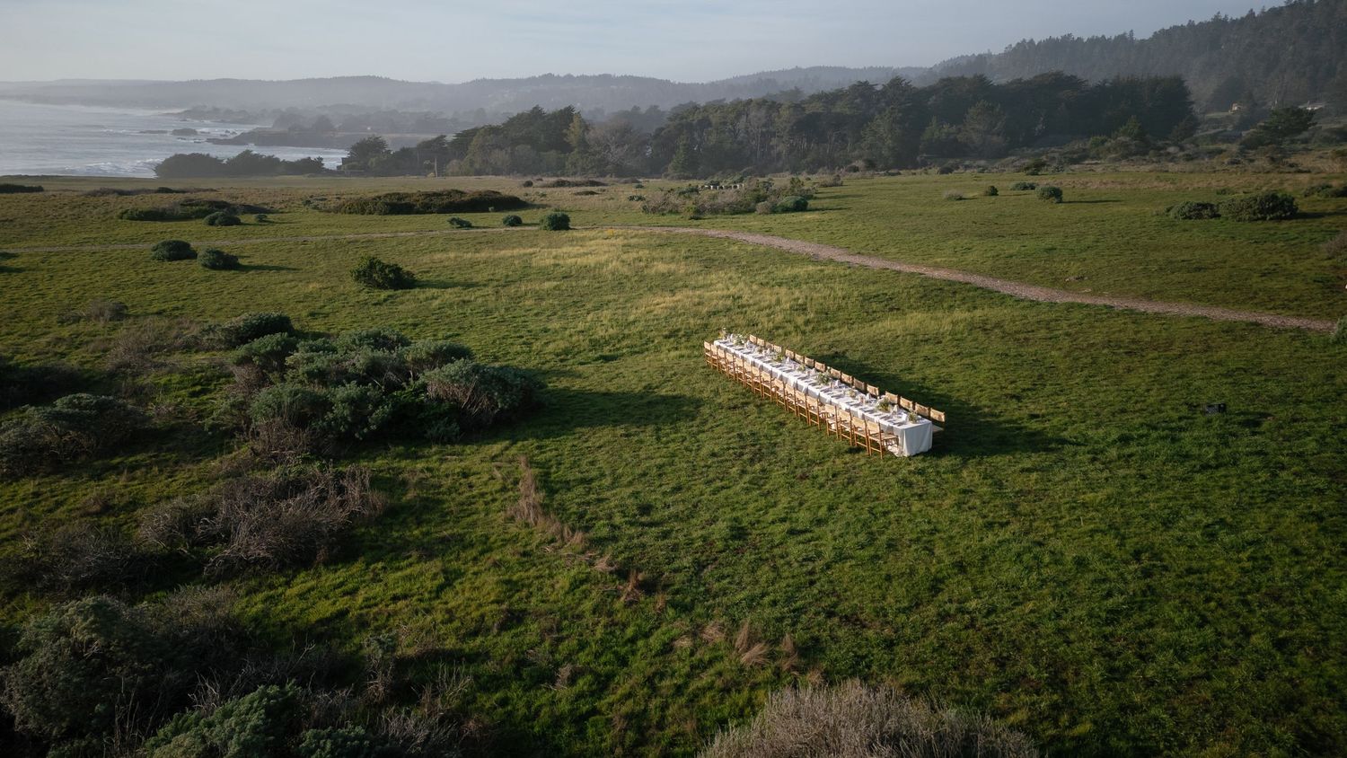 Aerial view of a lone shipping container resting in a green coastal field with rolling hills and ocean in the distance.