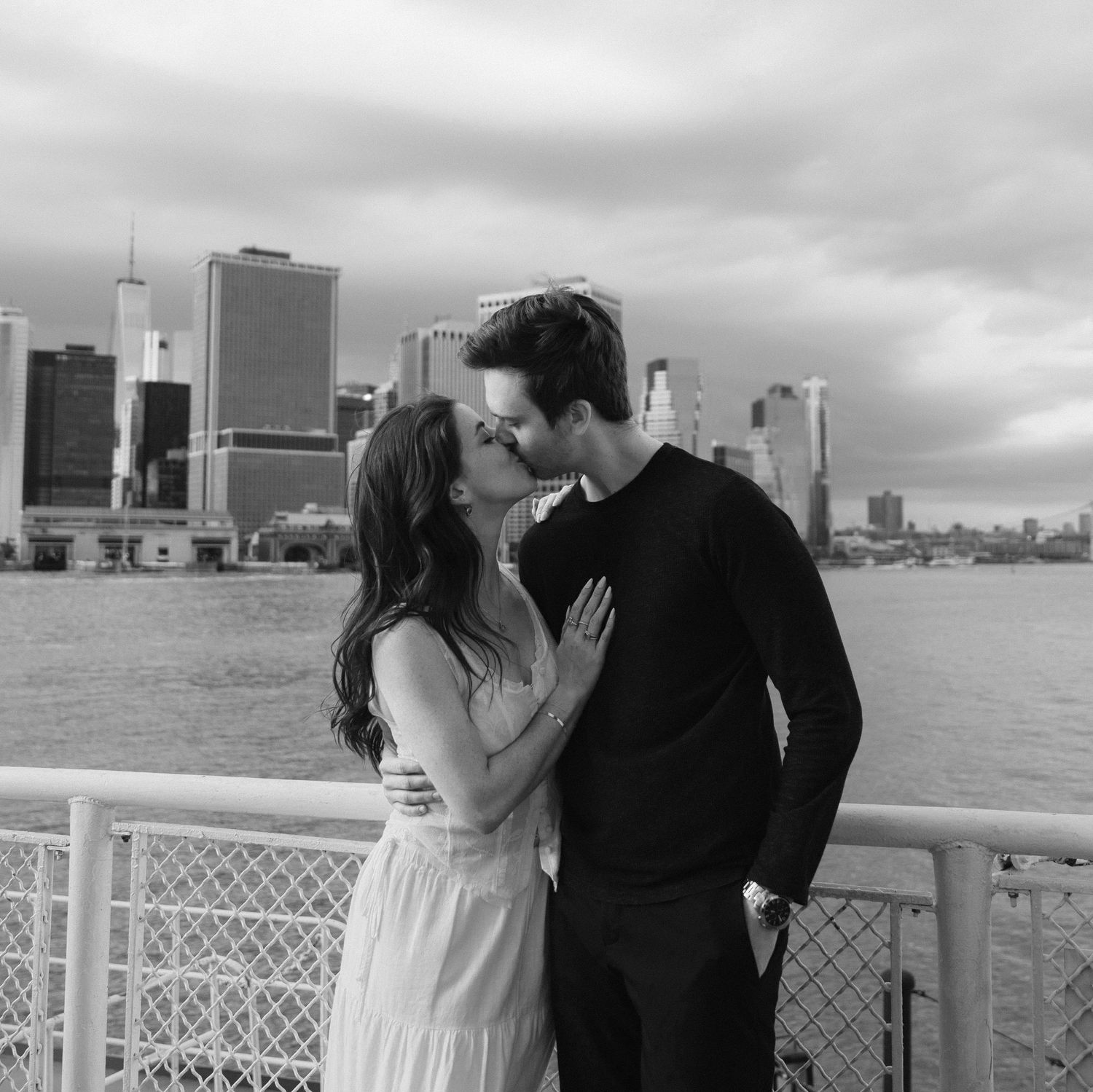 A couple shares an intimate moment on a waterfront with city buildings in the background.