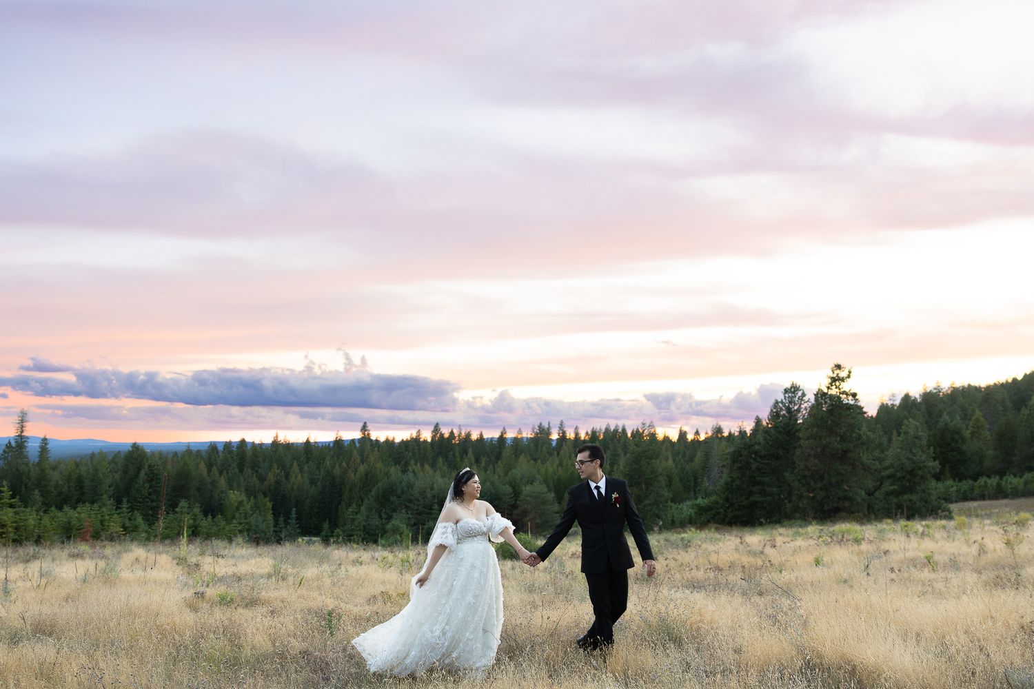 A couple in wedding attire walks through a scenic field at sunset with mountains and trees in the background.