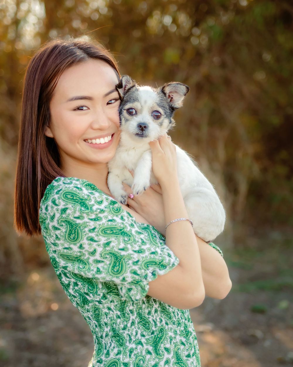 A senior girl wearing a green floral dress holds and cuddles a small white dog outdoors in natural lighting in Fort Worth