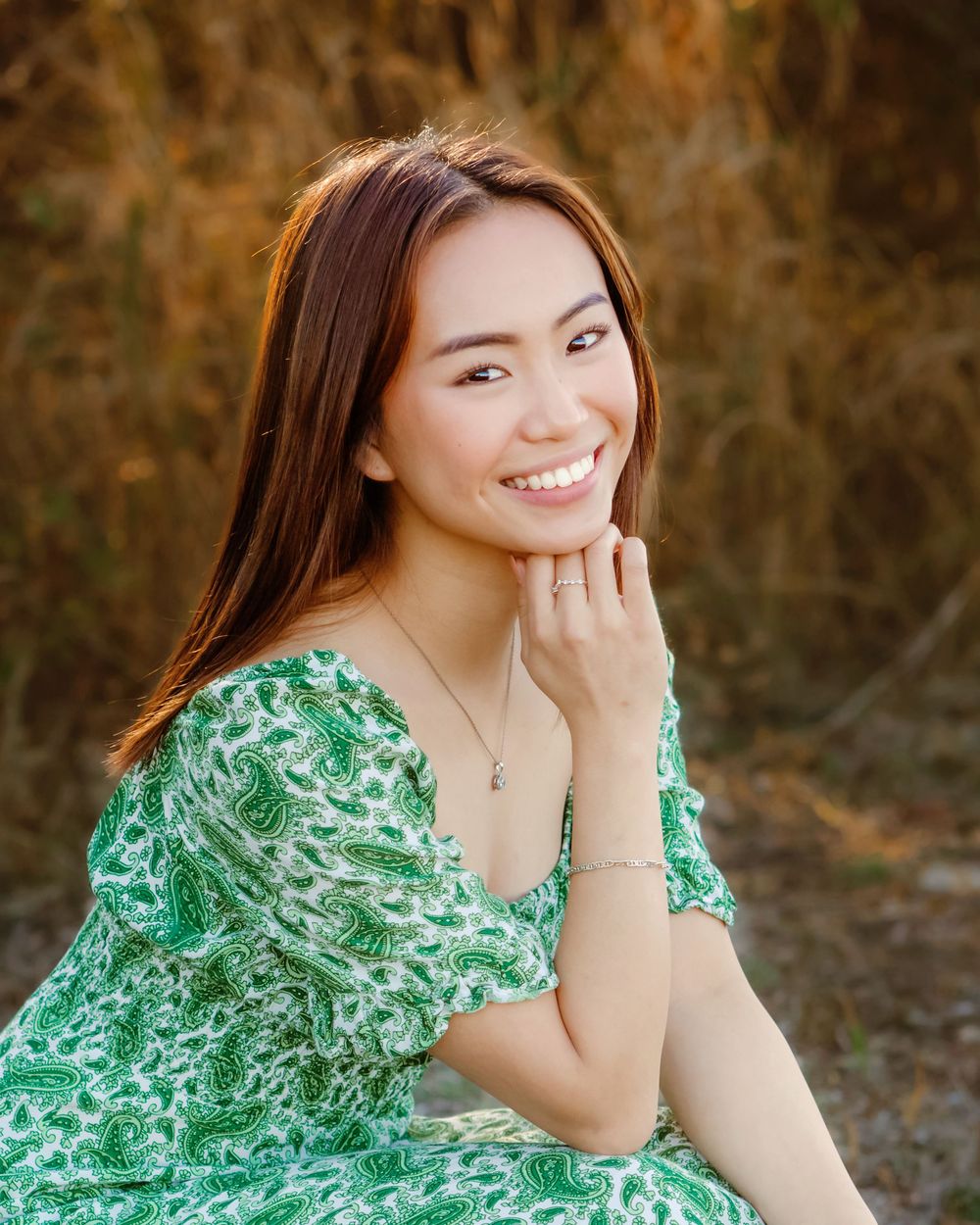 A senior girl in a green floral dress smiles warmly while posing outdoors against a natural brown background in Fort Worth