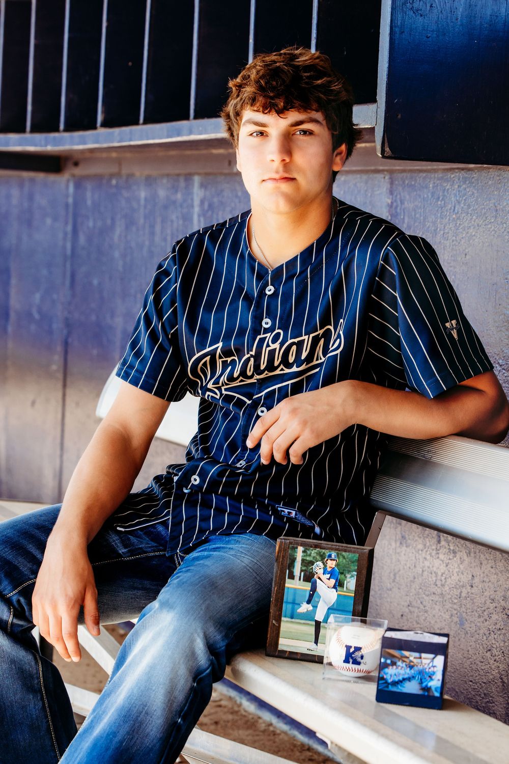 Teen boy in Indians pinstripe baseball jersey sits in dugout beside framed photo and memorabilia for senior portrait.