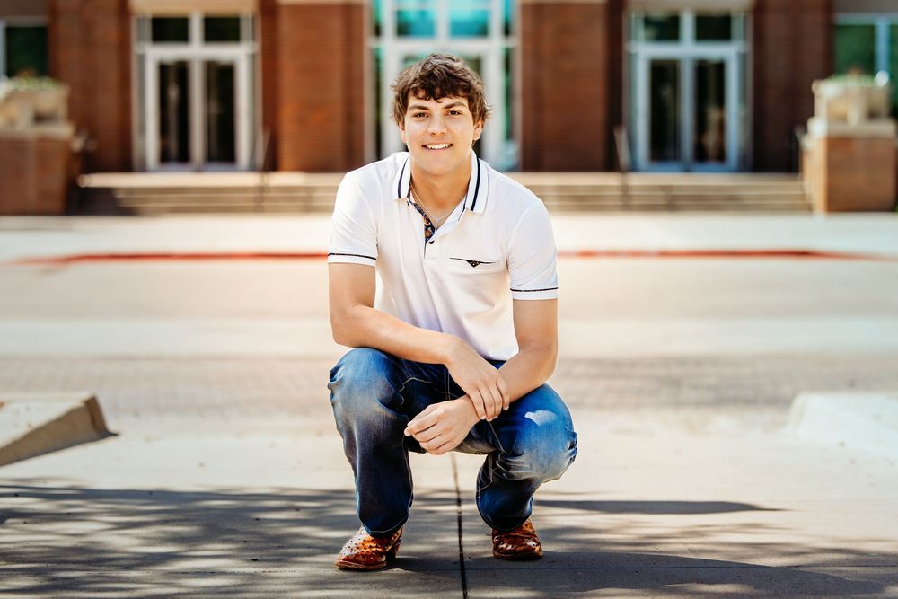 Young man in white polo and jeans crouching outdoors in front of a brick building on a sunny day.