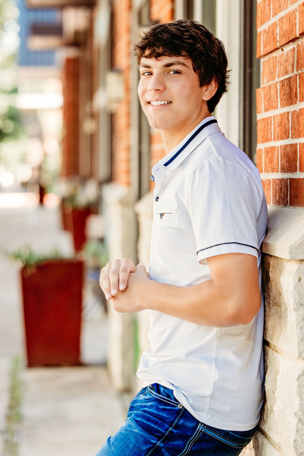 Young man in white polo shirt and jeans leaning against brick wall, smiling outdoors in urban setting.