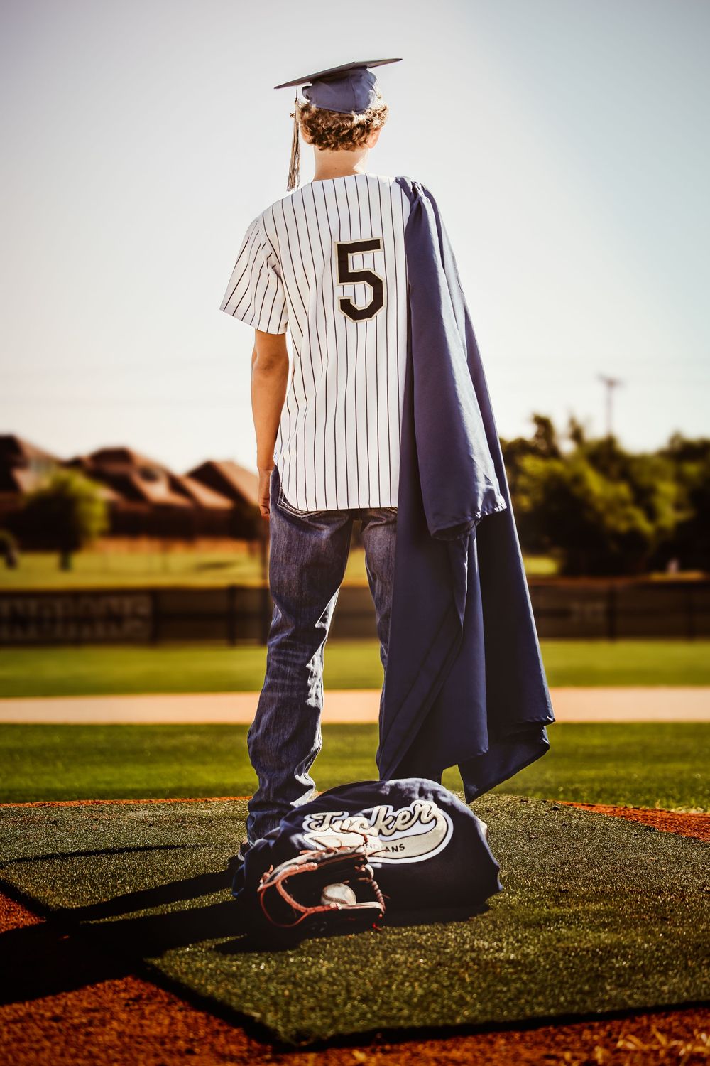 Graduate in cap and baseball jersey number 5 stands on pitching mound holding gown, celebrating dual achievement.