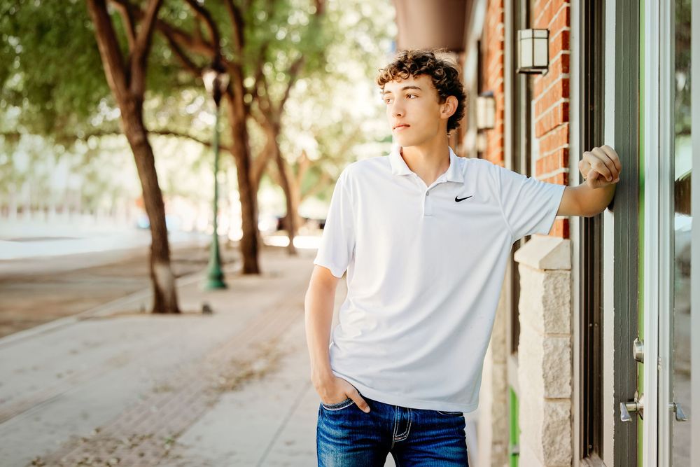 Young man in white Nike polo shirt leans against brick building on sunny urban sidewalk with trees in background.