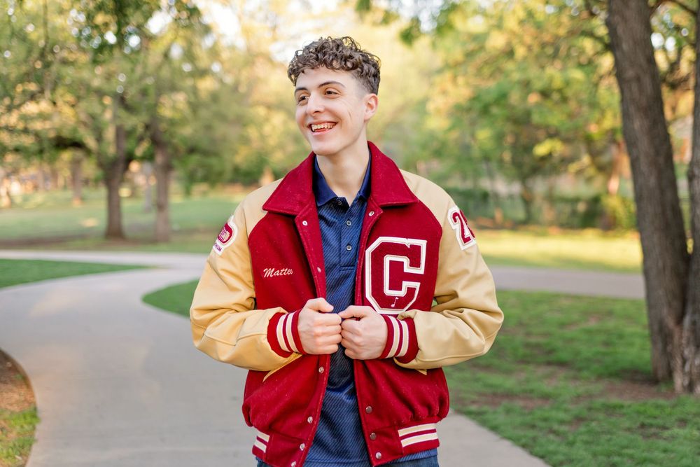 Senior boy in red varsity jacket poses outdoors during autumn photoshoot in park setting in Keller, TX