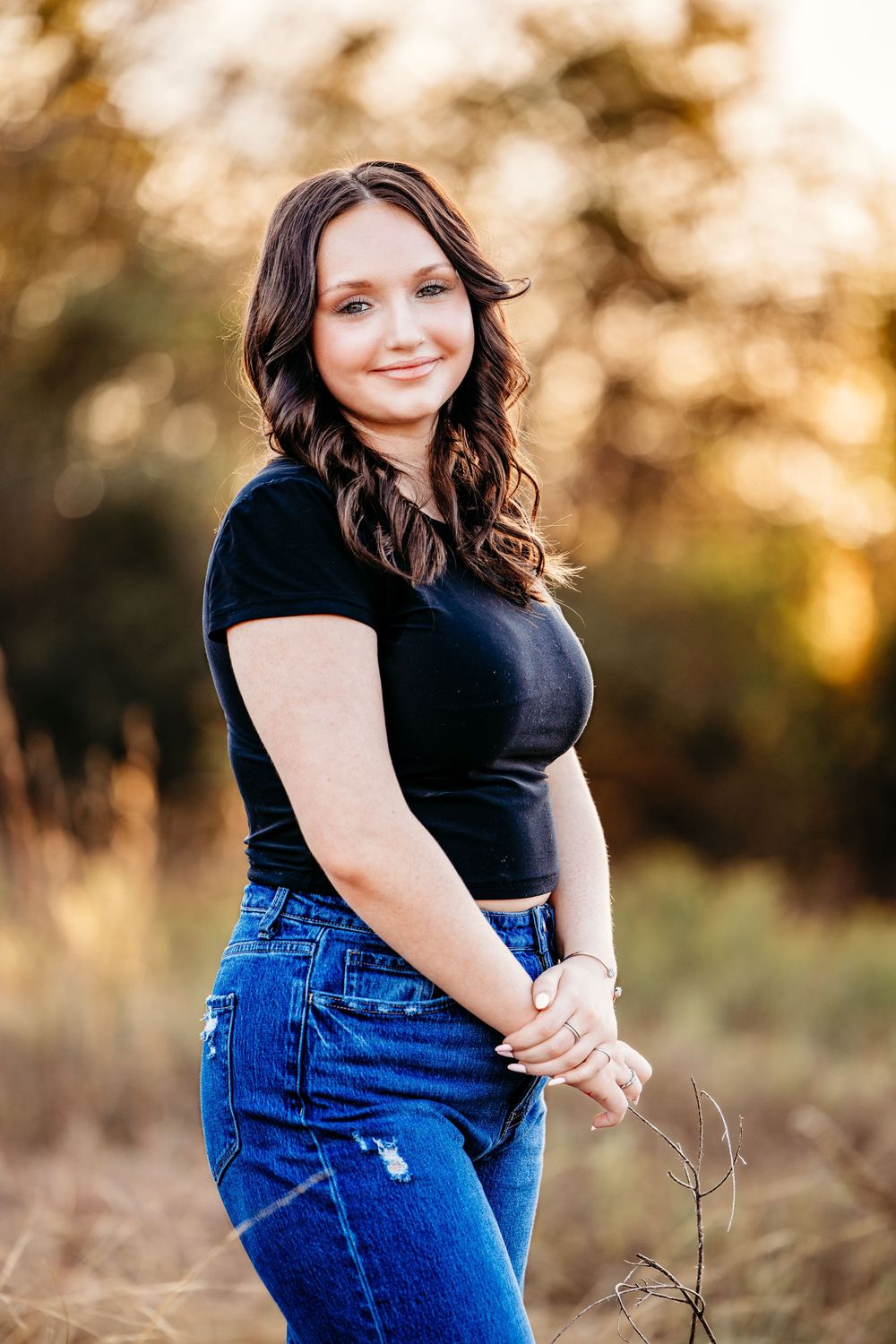 A senior girl in a black top and blue jeans poses outdoors during golden hour sunset in a natural setting in Fort Worth
