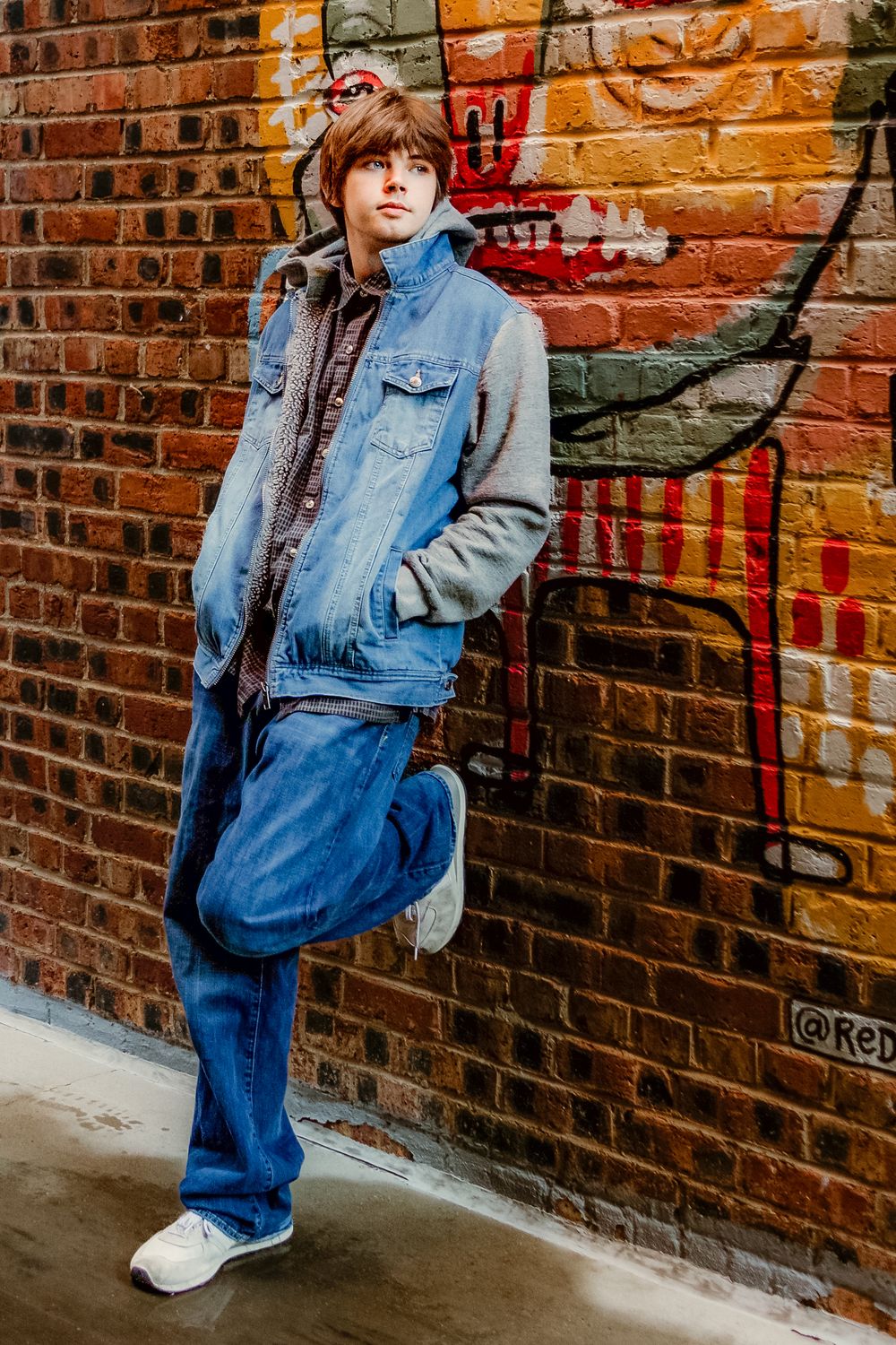 Senior boy in denim jacket and white sneakers poses against brick wall with graffiti in urban setting in Fort Worth