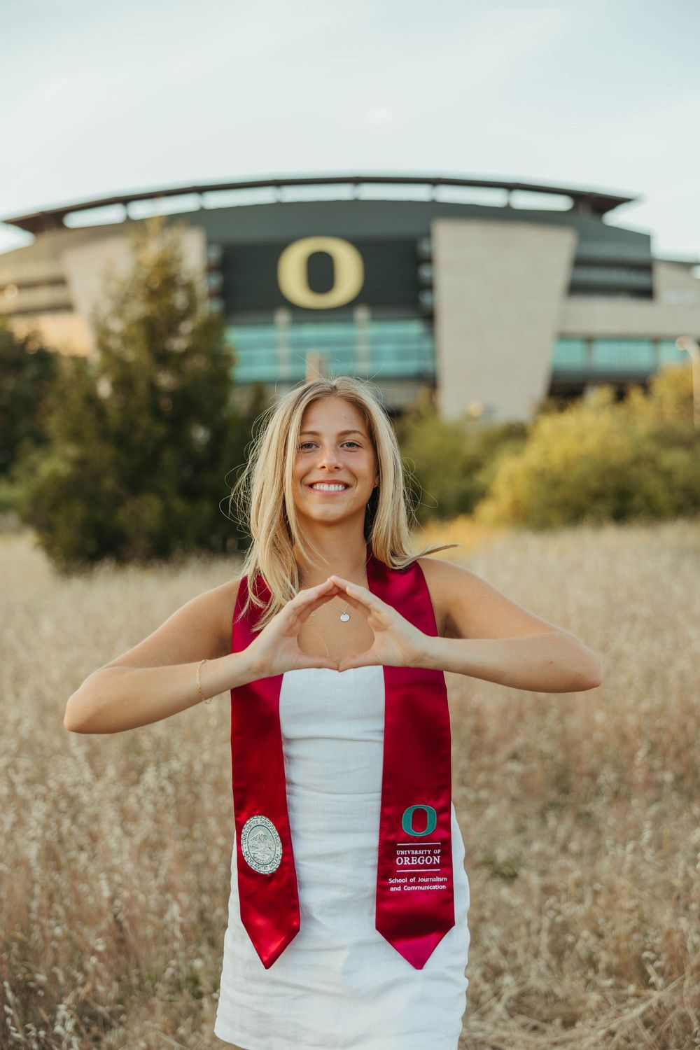 Graduate in red and white stole poses joyfully in field with university buildings in background.