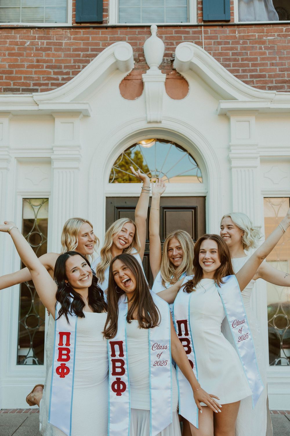 Group wearing white dresses and Greek letters celebrate together in front of a brick building with an ornate archway.