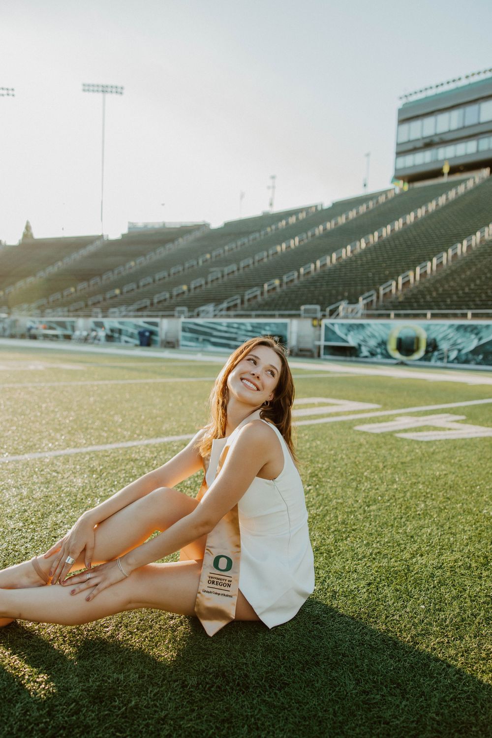 Person in white tank top sitting on football field turf with stadium seating and floodlights in background.
