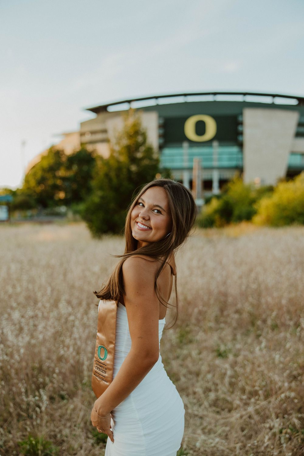 Woman in white dress poses in field with university stadium visible in background during golden hour.