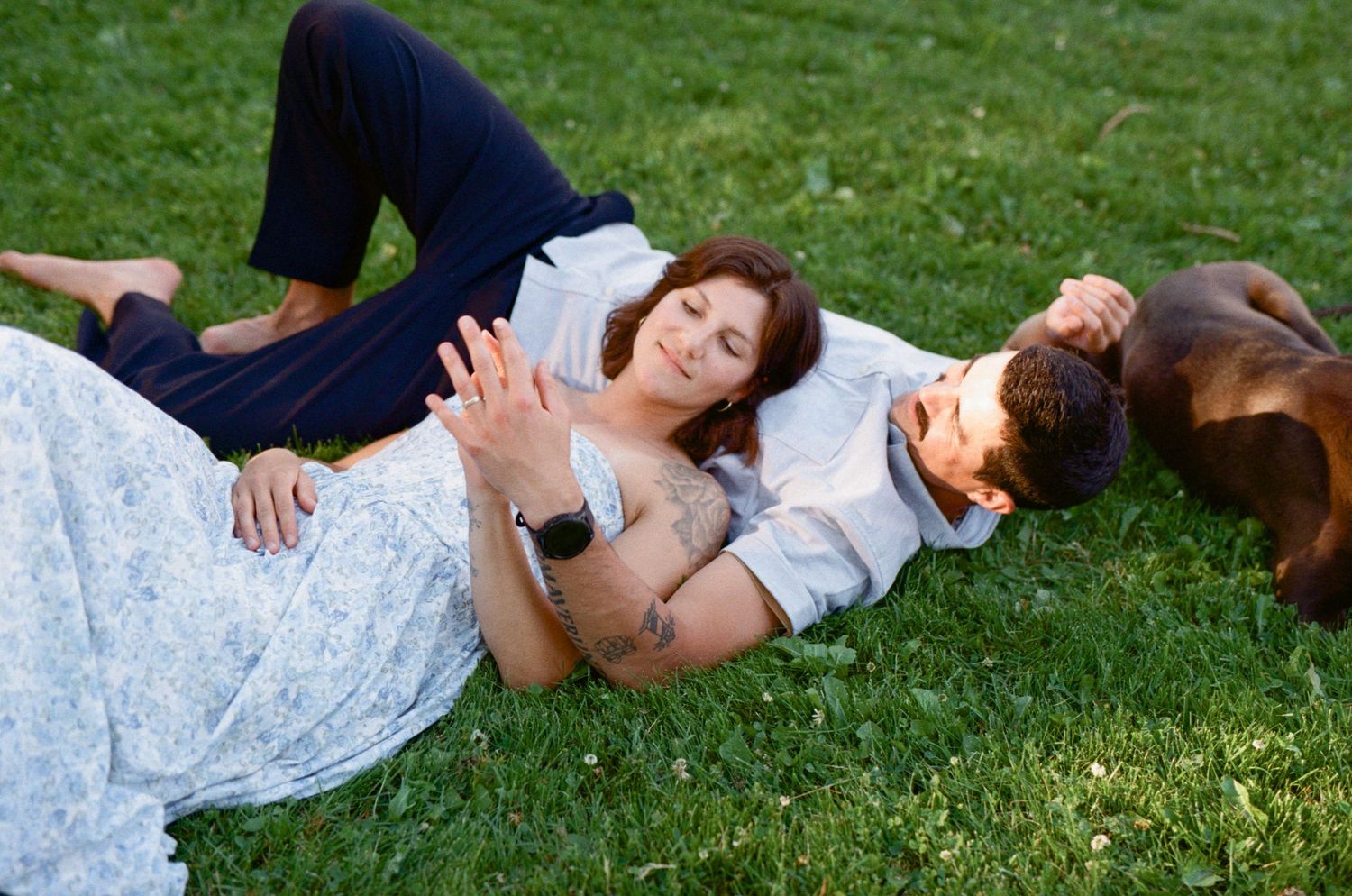 35 mm film photo of a woman laying on a man's chest in the grass, with their dog lying beside them in the evening light