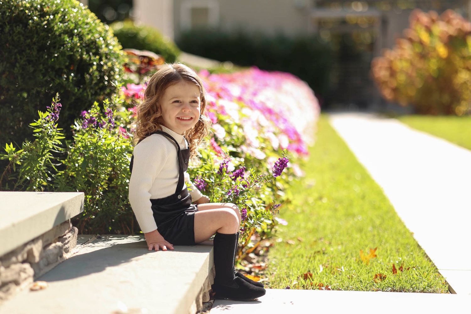 Smiling 3 year old boy sitting on steps next to flowers for outdoor Upsherin photography session in Jackson, NJ