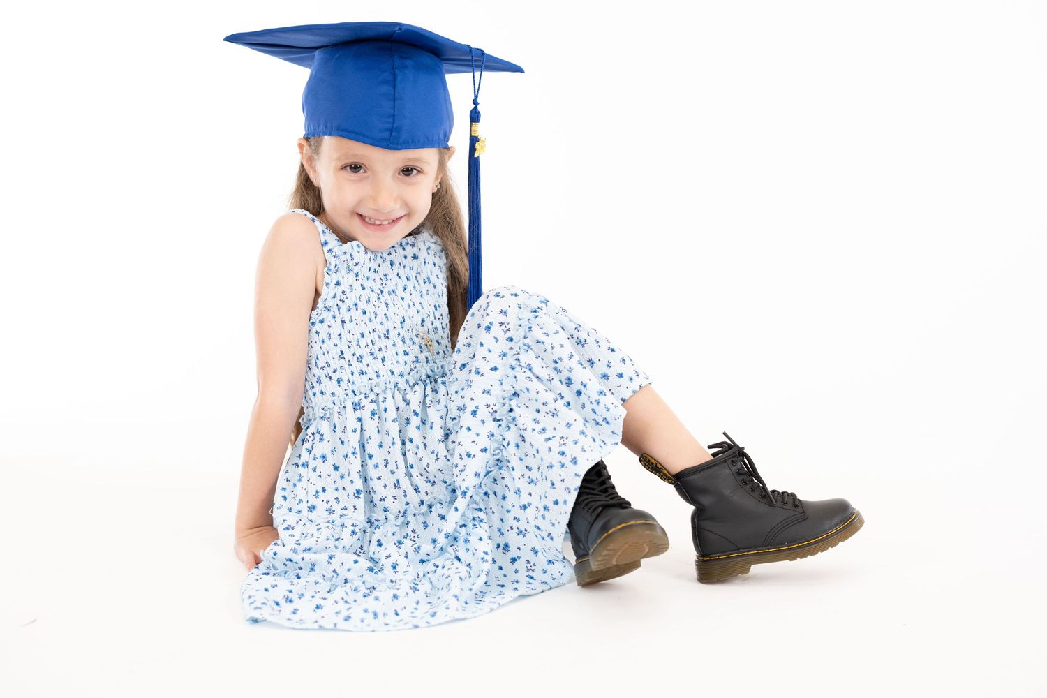 Child in blue graduation cap and floral dress sitting on white background.