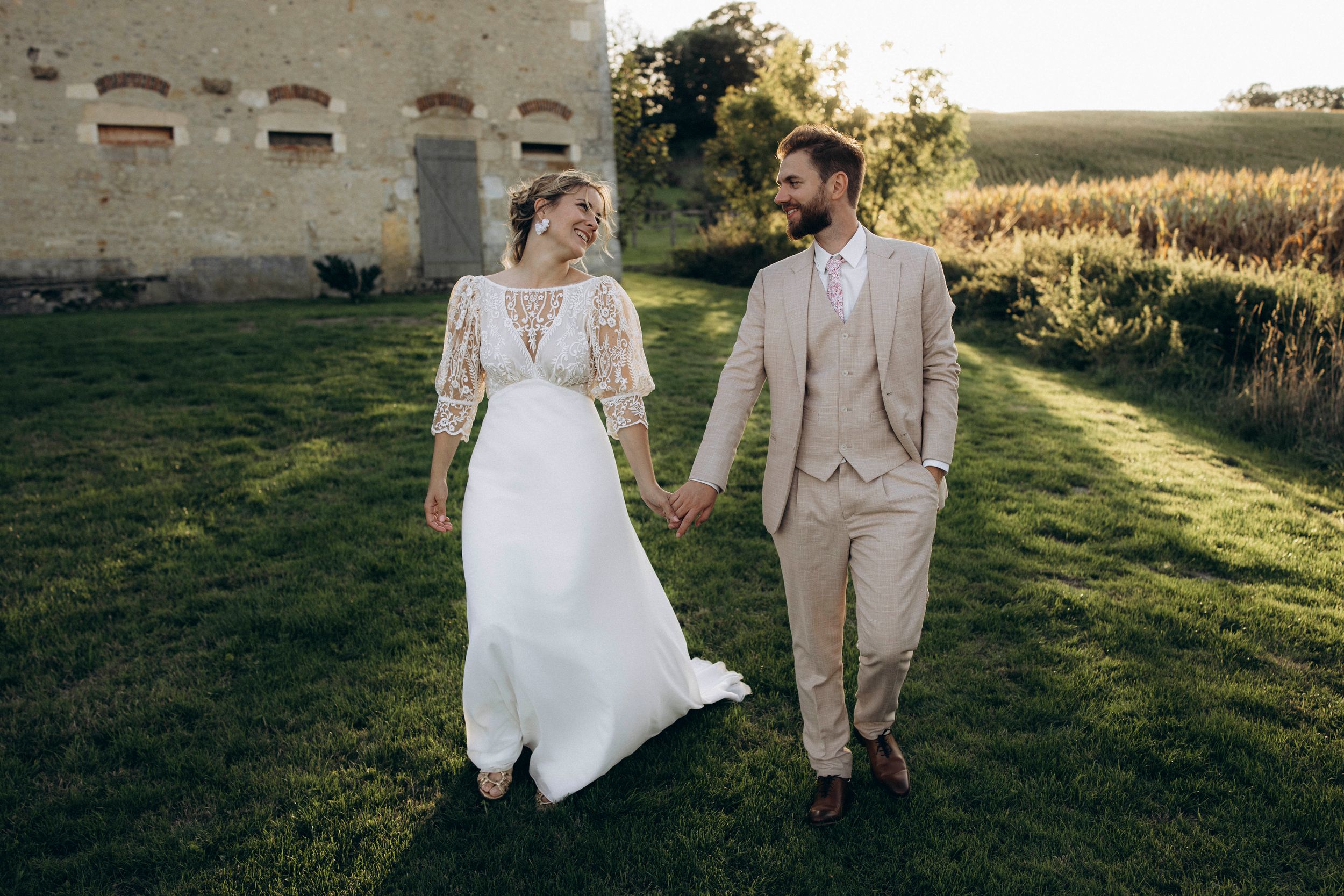 A couple in wedding attire walks hand in hand through a golden sunlit field during sunset.
