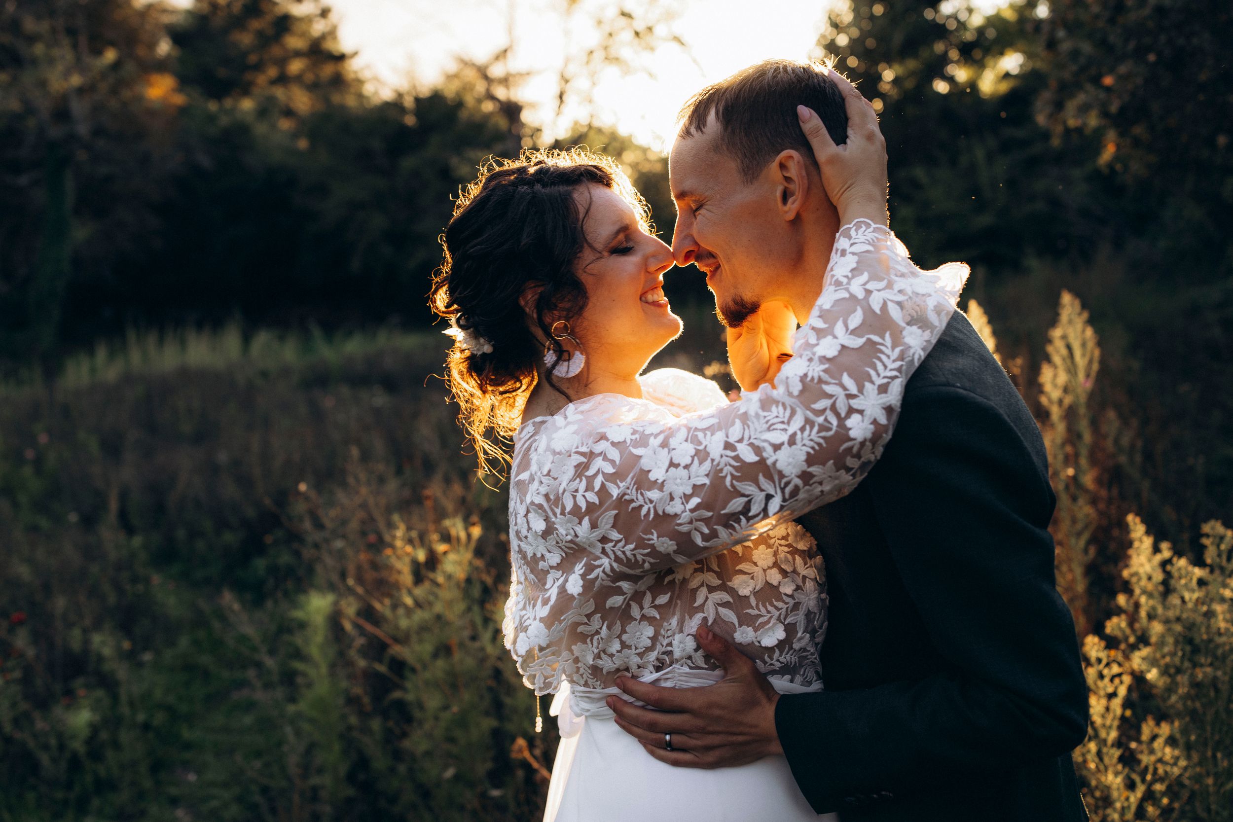 A couple in wedding attire shares an intimate moment at sunset in a wooded outdoor setting.