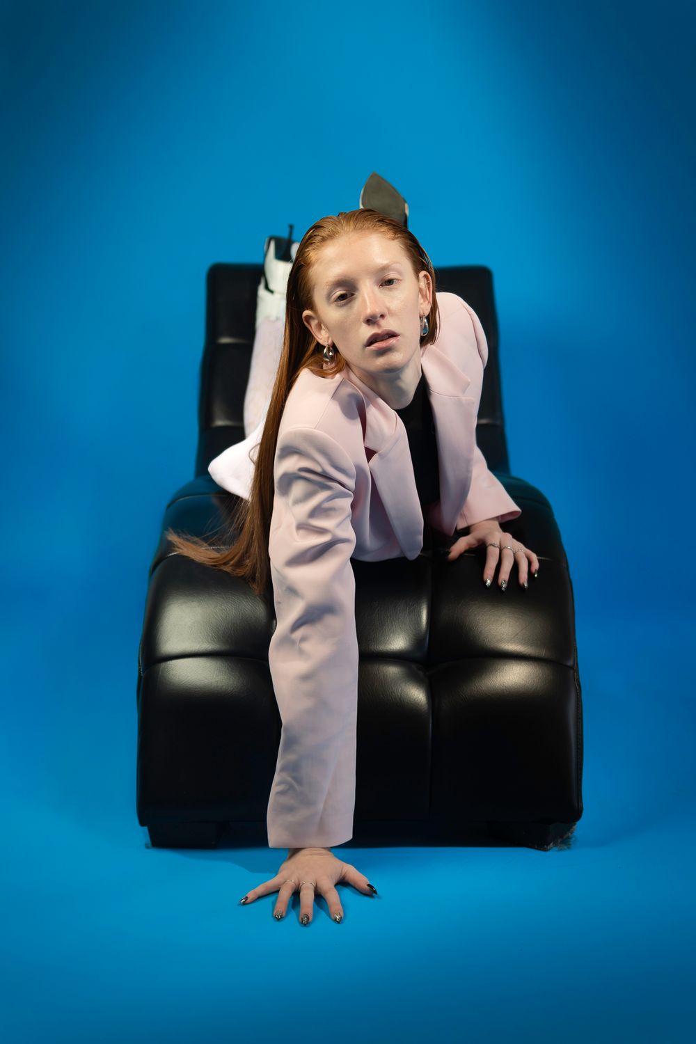 Figure in light colored clothing poses dramatically on black leather furniture against blue background.