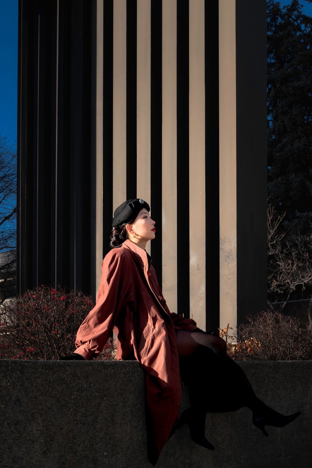 Artistic portrait against striped architectural columns in warm sunlight wearing red coat.