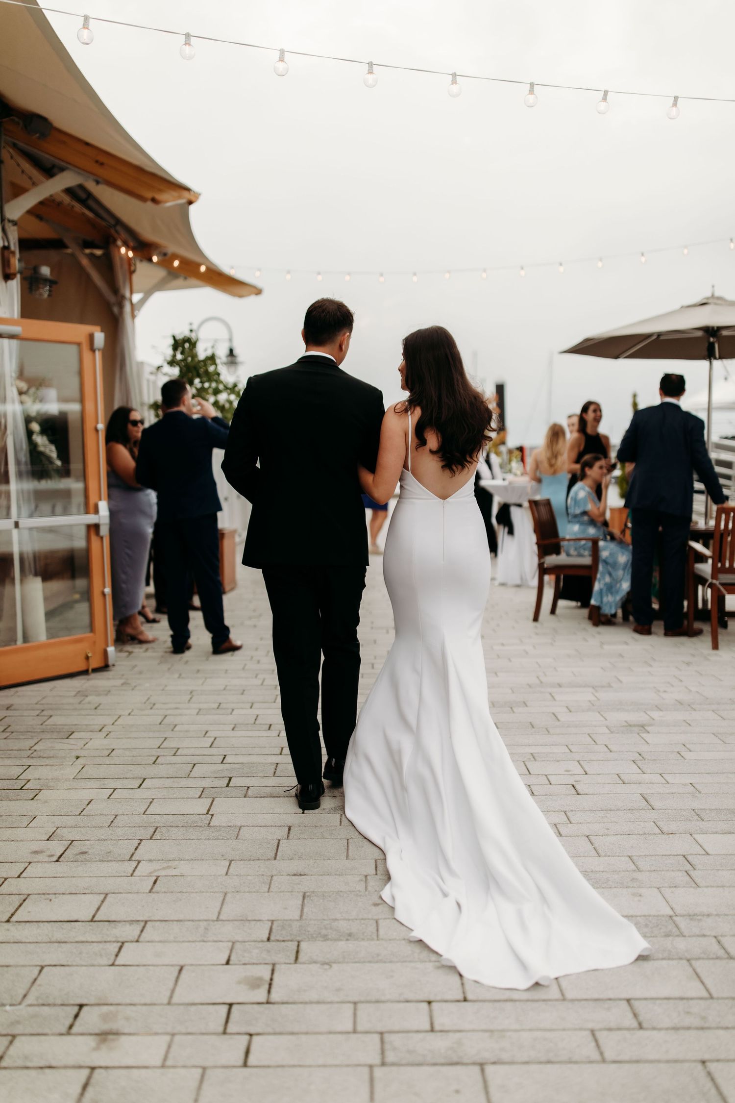 Wedding party walking through outdoor reception venue with string lights overhead.