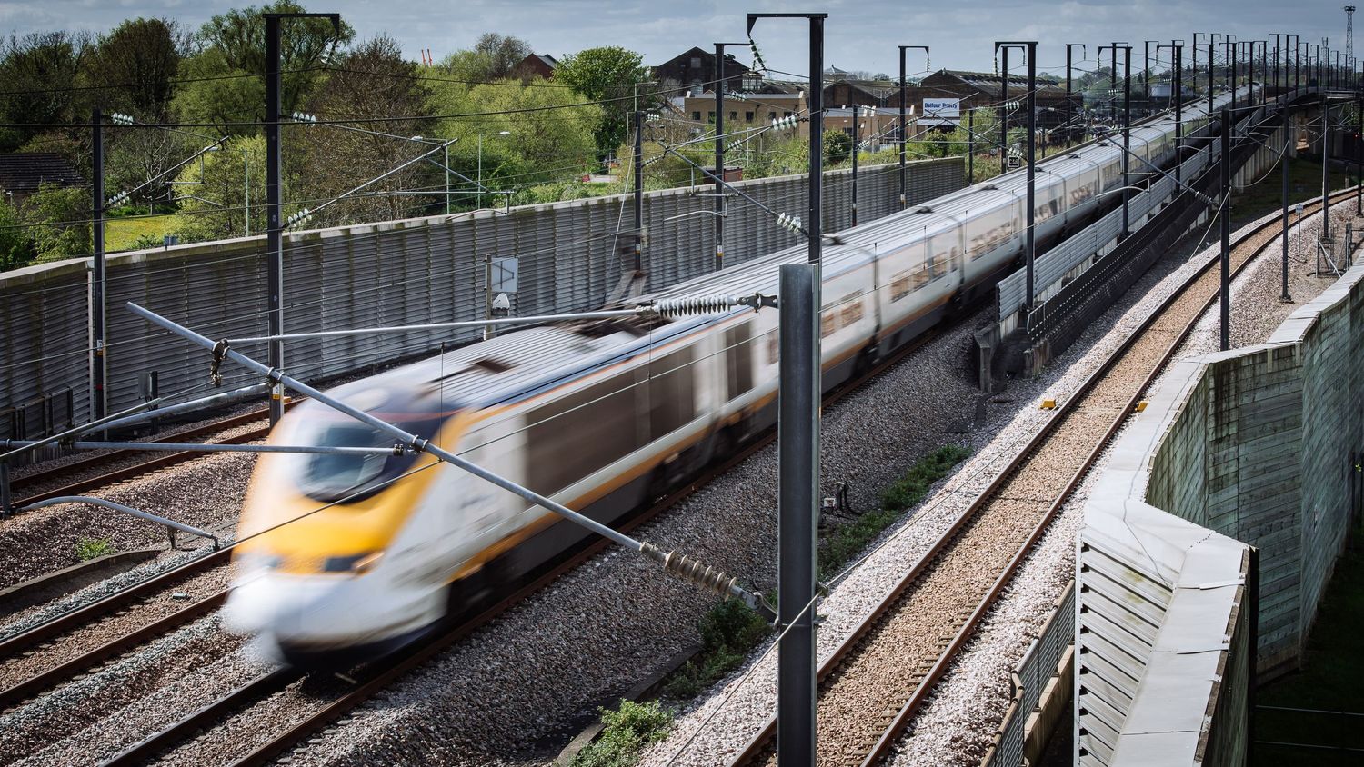 High-speed train passing through mountainous railway tracks with overhead power lines.