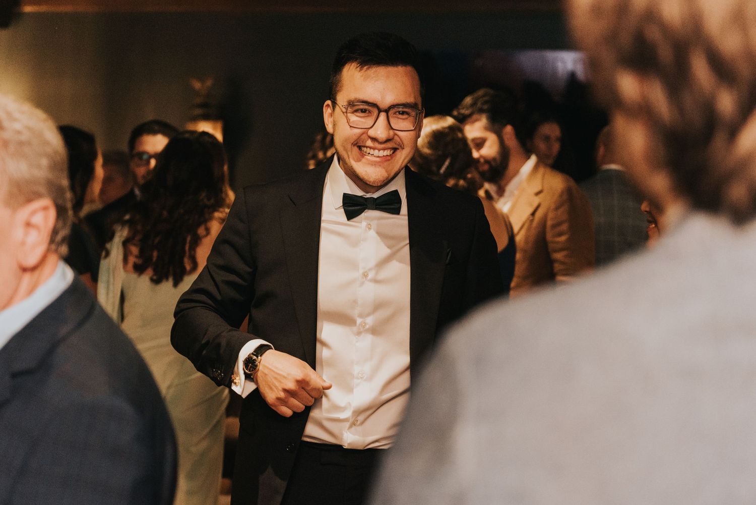 A guest in a black bow tie and glasses dances joyfully at an evening celebration.