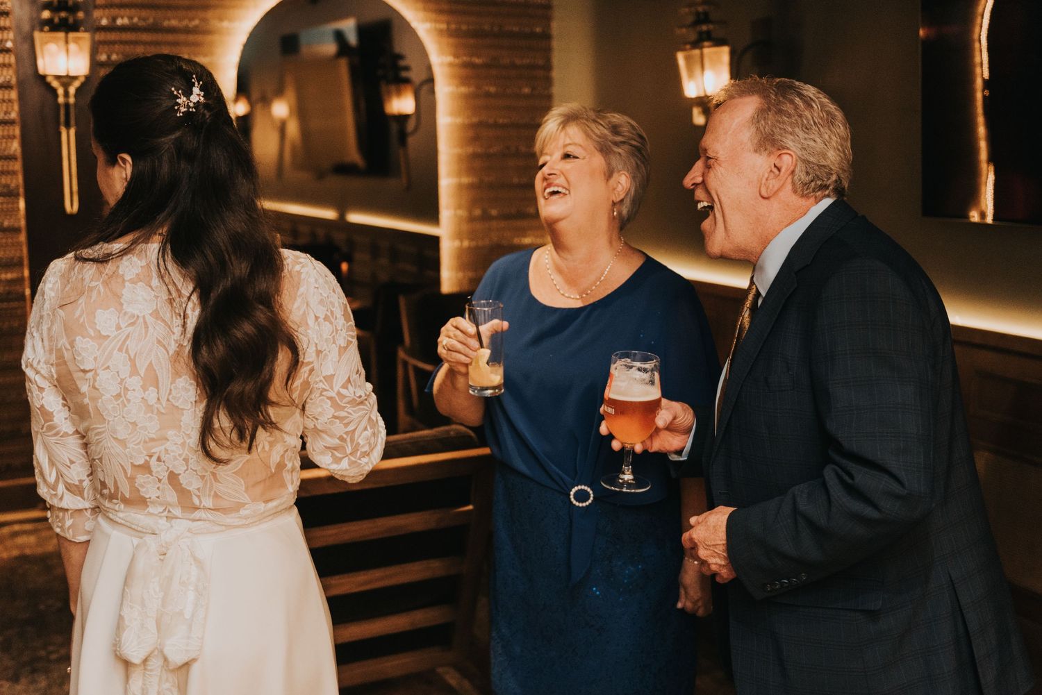 Three guests share drinks and laughter at an elegant evening reception with warm lighting.