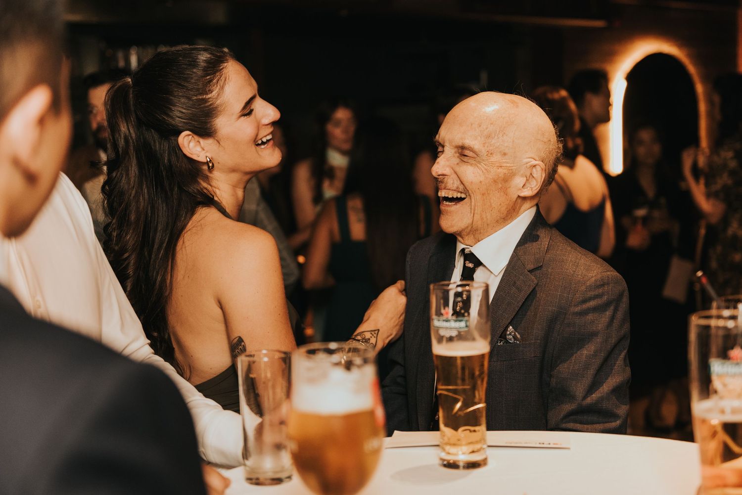A group of people socializing at a dimly lit evening wedding reception with drinks and laughter.