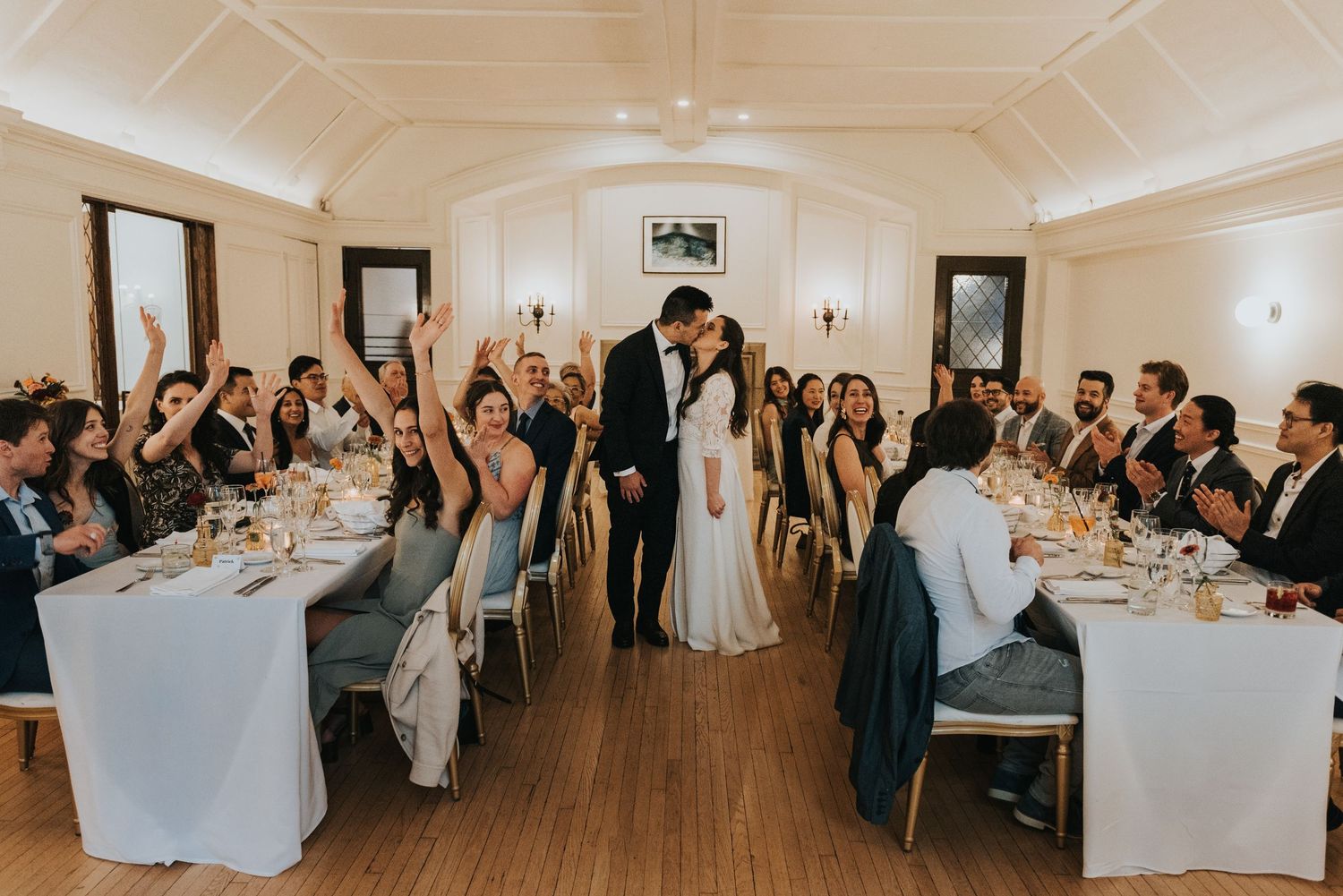 Panoramic view of an elegant wedding reception in a white-walled venue with guests seated at long dining tables.