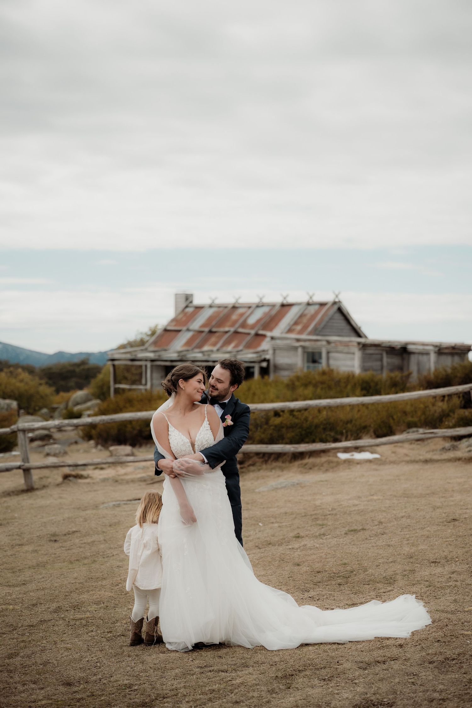 A couple in wedding attire embraces on a windswept hilltop with rustic buildings in the background at Craigs Hut