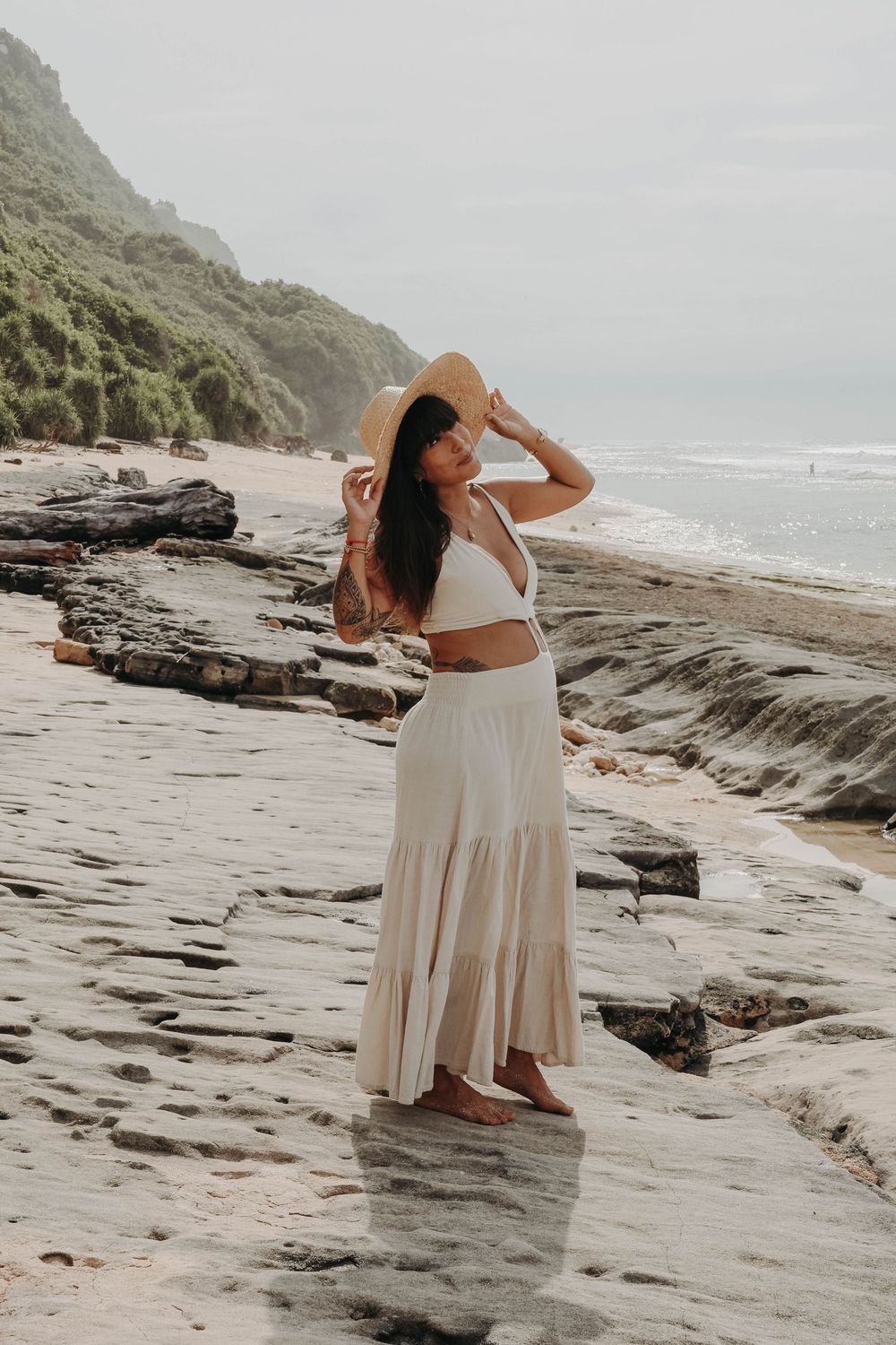 Pregnant woman in white crop top and maxi skirt holding straw hat on rocky beach.