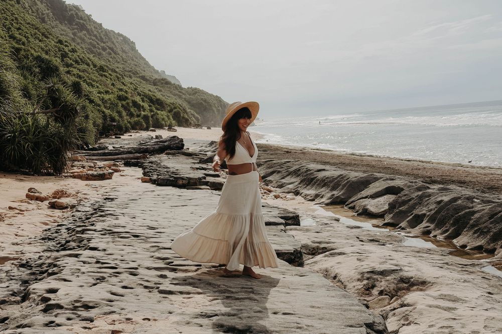 Pregnant woman in boho white outfit and straw hat walking on scenic rocky beach.