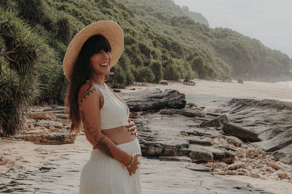 Smiling pregnant woman in white crop top and hat posing on rocky tropical beach.