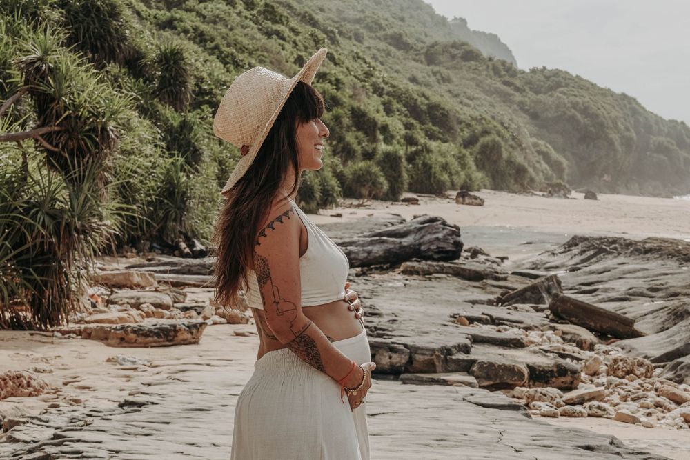Side profile of tattooed pregnant woman in white outfit on rocky beach with greenery.