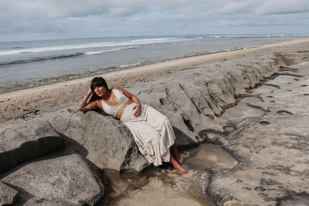 Pregnant woman in white maxi skirt reclining on flat rocks at the beach.