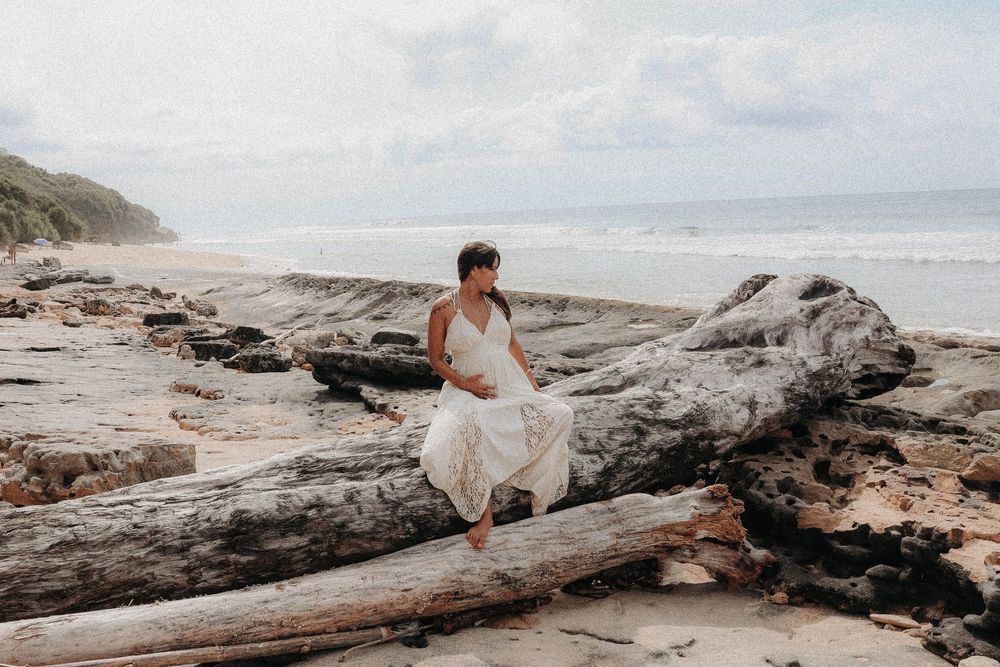 Woman in white dress sitting on driftwood logs on a tropical beach with ocean waves in background.