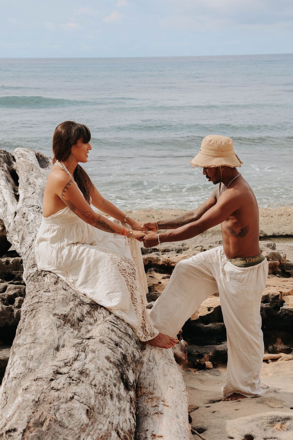 Couple holding hands on driftwood log at rocky beach with turquoise ocean in background.