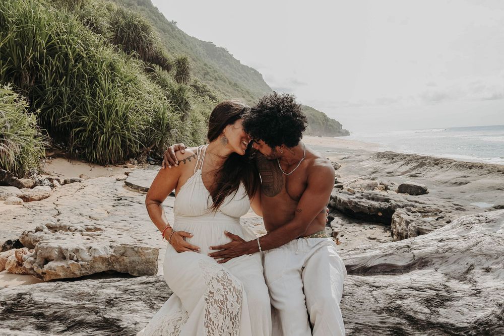 Happy couple sharing a tender moment on rocky beach with lush green hillside behind them.