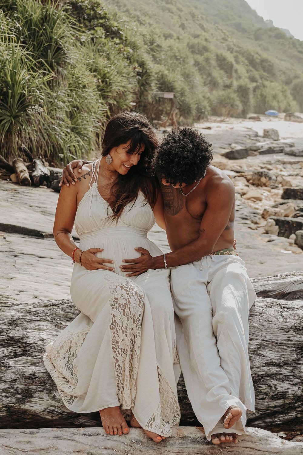 Couple sitting on coastal rocks, man embracing pregnant woman in white lace maternity dress.