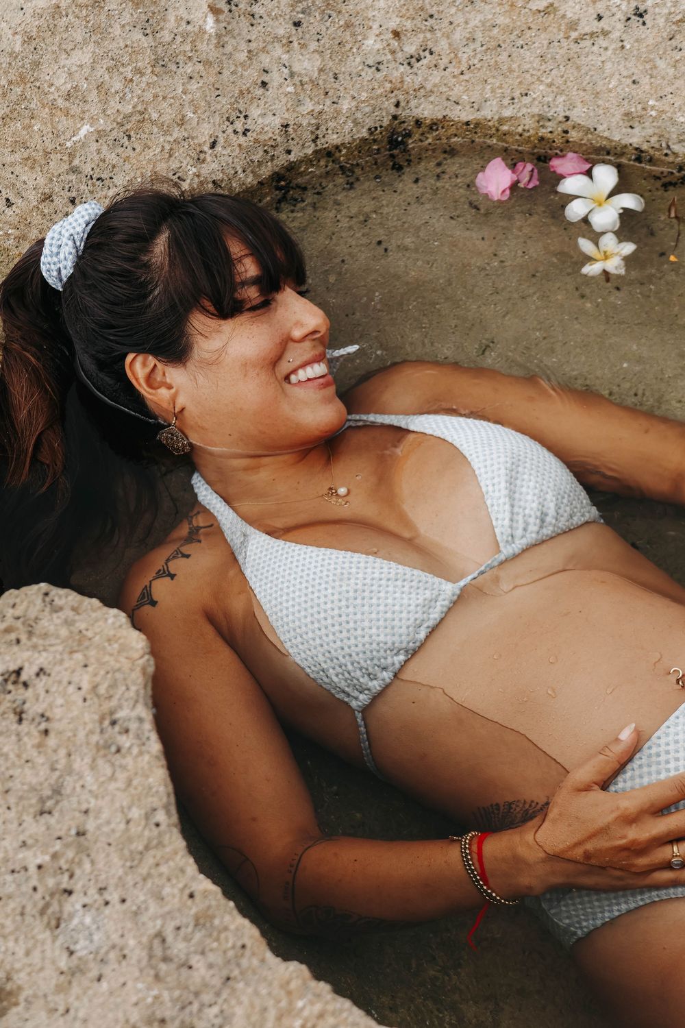 Smiling pregnant woman in white bikini relaxing in a rock pool with tropical plumeria flowers nearby.