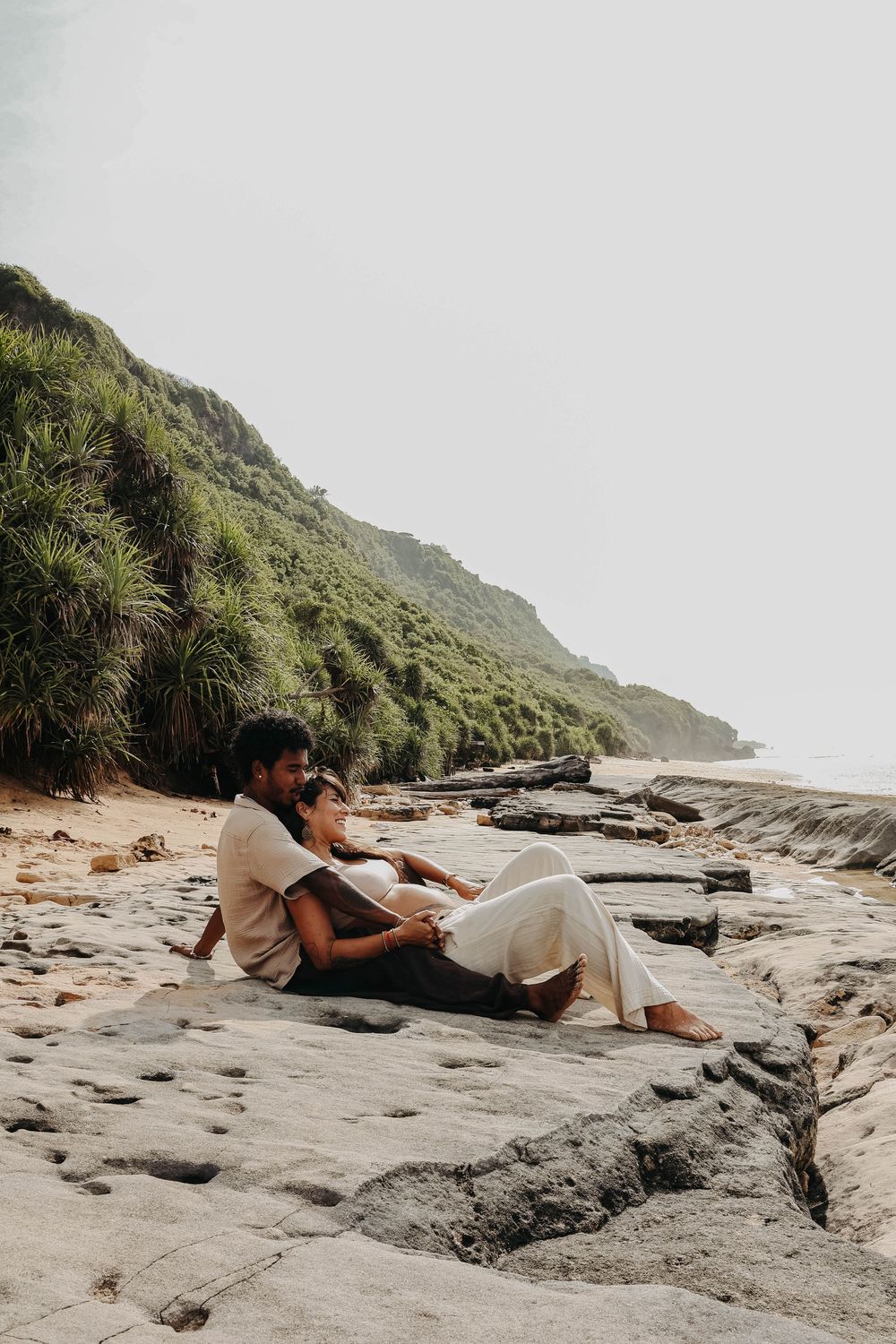 Couple sitting together on rocky beach, embracing with lush green hillside and ocean in background.