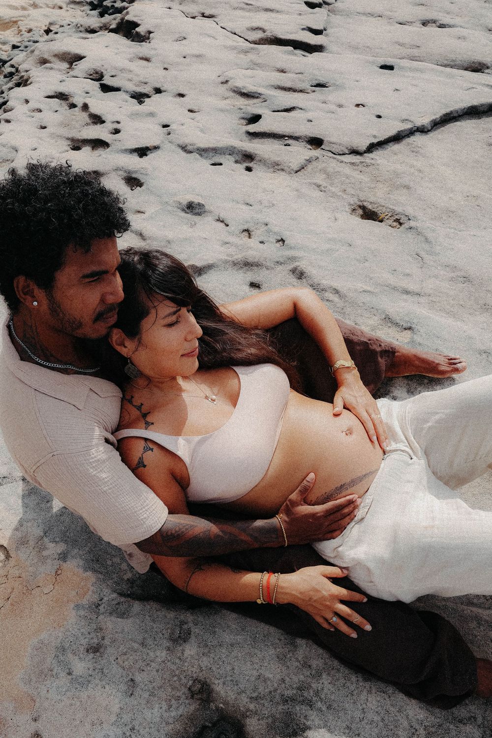 Couple reclining on beach rocks, man lovingly embracing pregnant woman with visible baby bump.