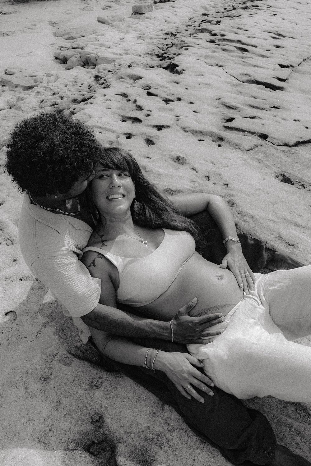 Black and white photo of couple on beach, man holding pregnant woman from behind on rocky shore.