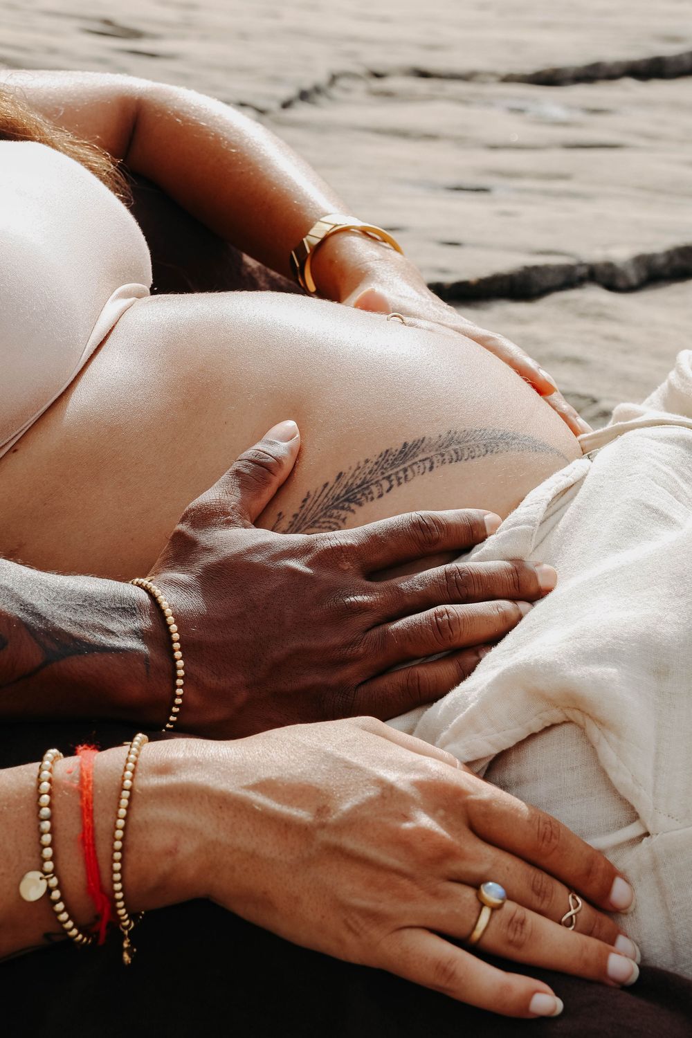 Close-up of couple's hands gently cradling pregnant belly with feather tattoo on rocky beach.