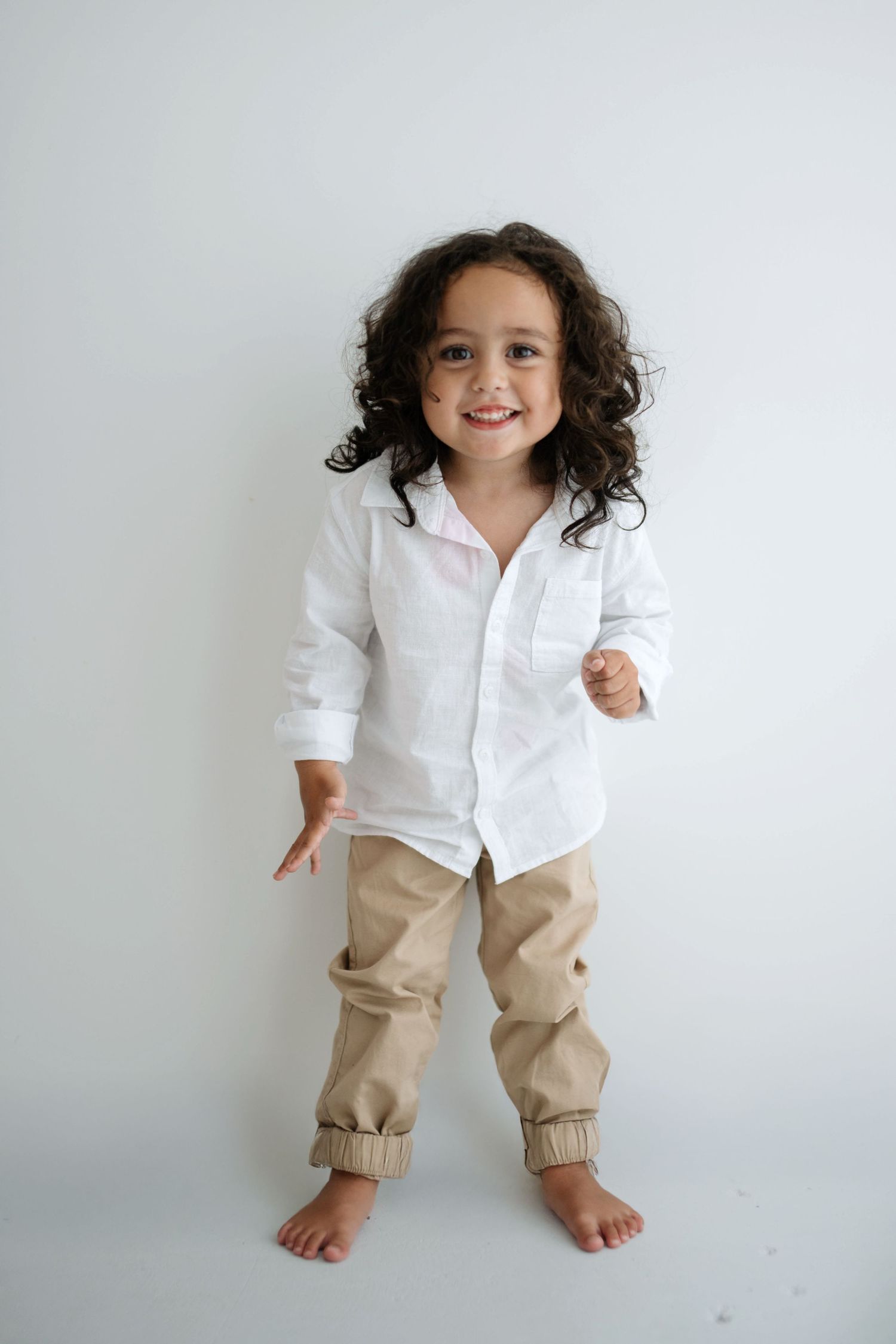 A child in a white button-down shirt and khaki joggers poses playfully against a light background.