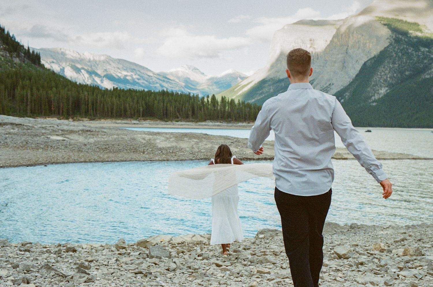 35 mm film photo of groom walking towards bride, who is twirling on the shore of Lake Minnewanka surrounded by mountains.