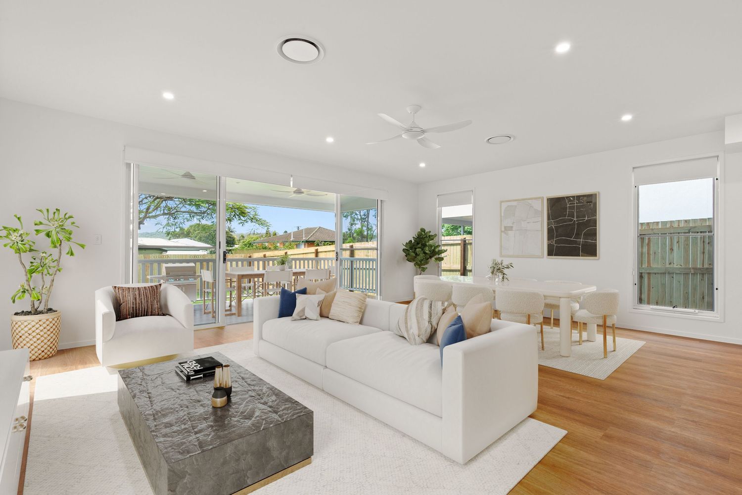 Sun-filled white living room with timber floors, large windows, white sofa, grey ottoman, and outdoor garden views.