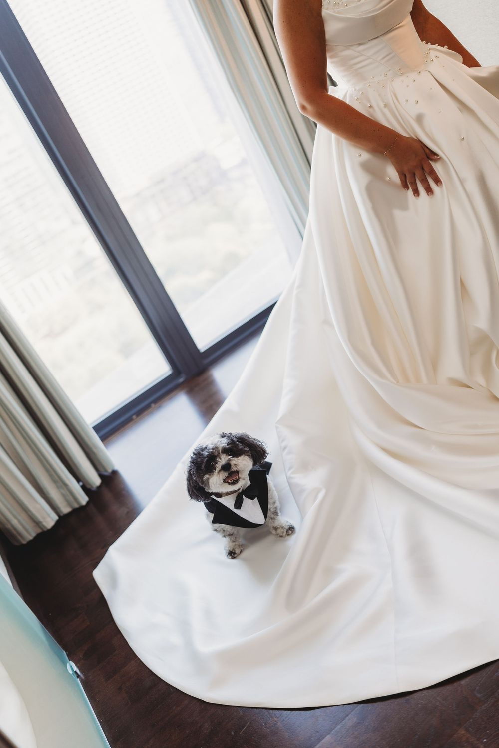 A flowing white wedding dress train cascades across dark hardwood floors in a bright room with natural window light.